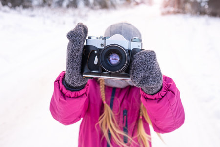 Close-up of slr film camera in raised hands of girl on winter walk. Child taking a photo. Day of photographer. Hobby, photography school. Portrait lens looking at camera.の写真素材