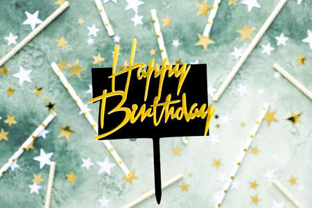 Golden lettering happy birthday on black plate with pen on blurred green background with stars and straws for cocktail. Festive birthday card, congratulation.の写真素材