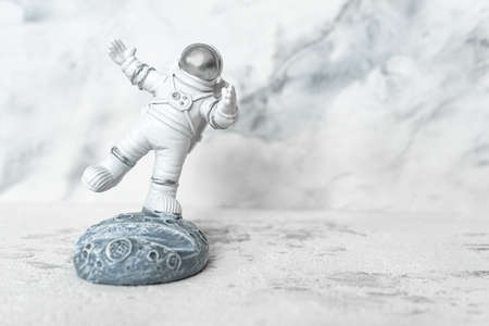 Miniature astronaut balancing on moon in spacesuit waving his hand on white. Phone stand in form of cosmonaut. World Day of Aviation and Cosmonautics. Science fiction, science and space exploration.の写真素材
