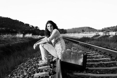 Beautiful brunette girl with suitcase sitting on railroad. Black and white photo.の写真素材