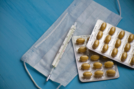 Pills, thermometer, medical face mask on a blue background, top view. First aid, care during...