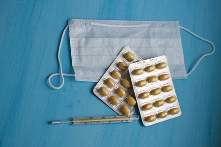Pills, thermometer, medical face mask on a blue background, top view. First aid, care during...