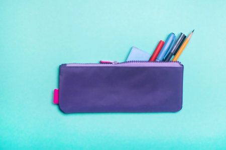 Fashionable violet pencil case with multicolored felt-tip pens, pencils and pens on green...