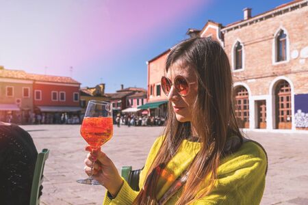 Young woman in yellow jacker with glass of aperol sitting on square in sunny day. Burano, Venice,...