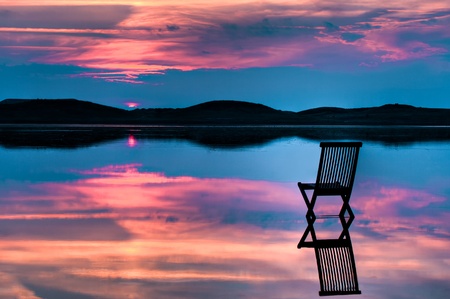 Scenic view of sunset over inlet and hills with a chair in the calm water, with reflections of sunset and chair. Symbolizing peace, loneliness or emptynessの写真素材