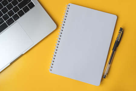 Modern office desk table with laptop and notepad with pen on yellow background. Bright backdrop with blank notebook page for text in the middle. Top view, flat layの写真素材