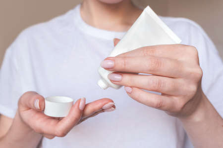 Female hands with natural pink manicure holding blank cosmetic tube. Woman in white holds beauty product. Concept of body care and skin protection. Moisturizing lotion closeup. Facial skincareの写真素材