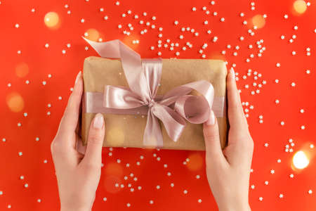 Nice female hands holding gift box wrapped in kraft paper with pink ribbon on festive red background with many snowflakes. Xmas and New Year postcard design. Giving love and warmth, Christmas conceptの写真素材