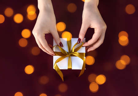 Nice female hands holding gift box wrapped in white paper with golden ribbon on festive dark...