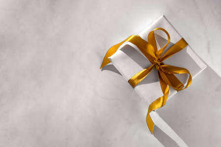 Gift box wrapped in white paper with a golden bow on festive light background. Copyspace for your text. Flat lay style. Christmas, New Year or birthday celebration concept. Web element for postcardの写真素材