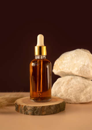 Transparent glass cosmetic dropper standing on wooden podium near stones and dry flowers on dark beige background. Beauty composition. Concept of natural and original skincare products presentation.の写真素材