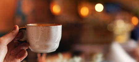 Closeup of female hand with natural manicure holding cozy ceramic white mug of tea or coffee. Web banner. Relax and comfort at home, cafe. Empty space for text on blurry background, backplateの写真素材