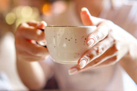 Closeup of female hand with natural manicure holding cozy ceramic white mug of tea or coffee.. Relax and comfort at home, cafe. Empty space for text on blurry background, backplateの写真素材