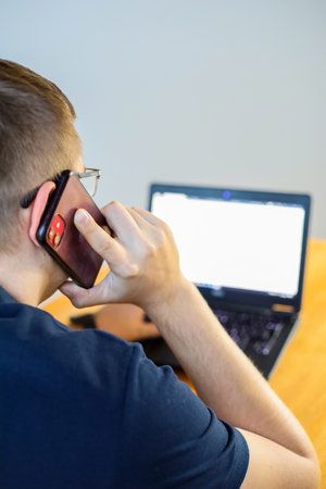 Young man using a mobile phone in front of a laptop computer.の写真素材