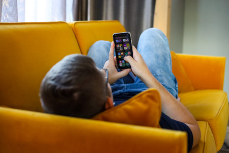 Young man sitting on sofa and using mobile phone in living room at homeの写真素材