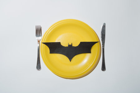 Black bat laid on yellow plate with fork and knife against white background. Minimal halloween concept. Flat lay.の写真素材