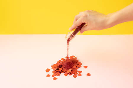 A beautiful female hand holds a test tube with sequins and spills over the red heart. Pink and yellow background.の写真素材