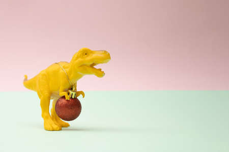 Yellow toy dinosaur tyrannosaurus rex with new year Christmas red baulbe around nack with golden rope on a pink and mint pastel background.の写真素材