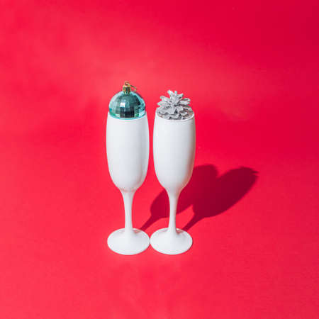 Two white champagne glasses with pine cone and light blue disco ball inside of them. Red pastel background. Minimal design.の写真素材