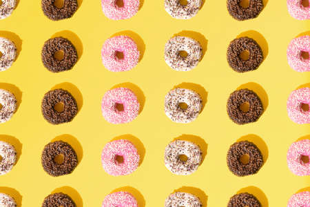 Arranged small ring donuts with white, pink and chocolate glaze. Colorful crumbs on a yellow pastel background. Pattern. Flat lay.の写真素材