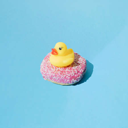 Small yellow rubber duck toy on donut with pink glaze and small white crumbs. Blue pastel...