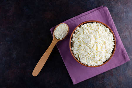 Cottage cheese and wooden spoon on a dark background. Fresh cottage cheese in a bowl. Clay plate with soft cheese on a linen napkin. Top view. Copy spaceの写真素材