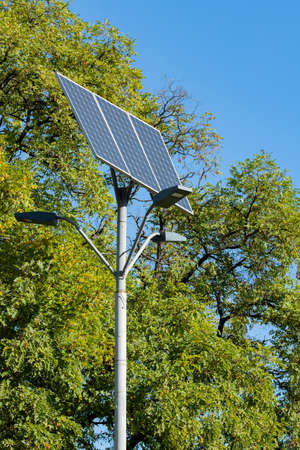 Alternative solar energy. Solar device with street lamp on background of blue sky and green tree....
