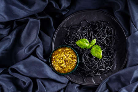 Black pasta, pesto sauce and basil on a dark background. Black spaghetti with cuttlefish ink on a black cloth. Pasta on black plate and sauce pesto. Copy space. Top viewの写真素材