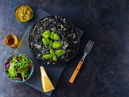 Black pasta with parmesan cheese, basil and pesto sauce. Black spaghetti with cuttlefish ink on a dark background. Pasta with grated cheese and fresh greens mix salad. Top view. Copy spaceの写真素材
