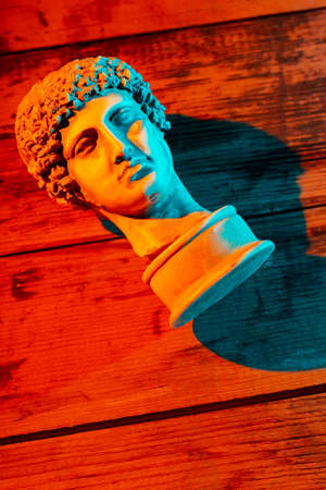 Gypsum copy of ancient statue Hermes head in orange turquoise neon light. Ancient greek sculpture Hermes head on wooden boards. Minimal art fantasy concept. Copy spaceの写真素材