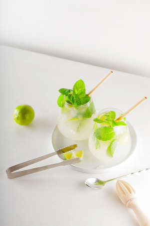 Two mojito glasses with straws. Mojito cocktail with lime and mint on white table. Summer refreshing cocktail and bar accessories on a white plateの写真素材