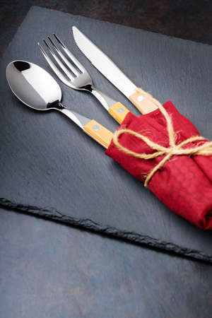 Cutlery with in a burgundy napkin on a dark background. Spoon, fork and knife on a slate board. Table setting. Top viewの写真素材