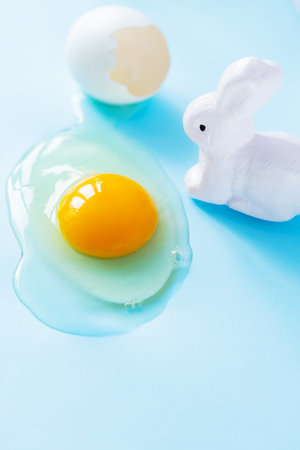 Broken chicken egg and white rabbit on blue background. Broken eggshell and egg yolk. easter conceptの写真素材