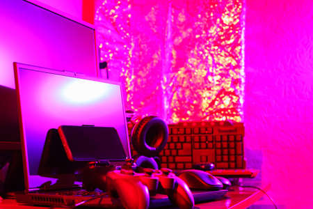 Gamer's workspace. Gamepads, computer mouse, headphones, keyboard and laptop on the table. Gaming accessories and computer equipment in neon light. Gambling addiction conceptの写真素材