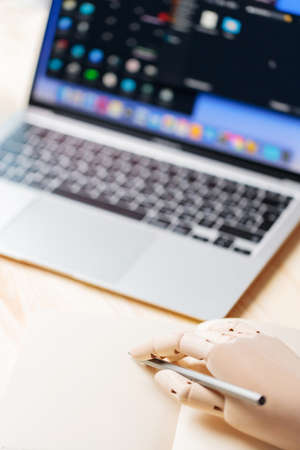 Robotic hand holding a pencil near a notebook. Robot arm works next to open laptop. Artificial...