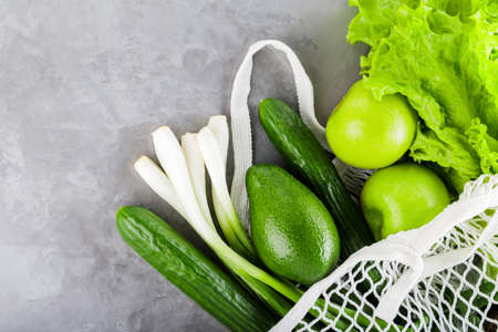 Reusable grocery net bag with fresh vegetables and fruits on gray concrete. Zero waste mesh bag with...