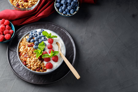Breakfast cereal granola with berries and greek yogurt on a dark background. Bowl with yogurt, granola and fresh berries on a black plate. Healthy breakfast with wooden spoonの写真素材