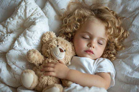 A charming little girl with blonde curly hair sleeps serenely on white bed linen, gently hugging a soft teddy bear, the concept of sleep and rest, healthy lifestyle and developmentの素材