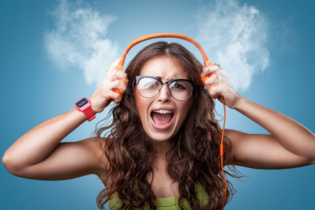 Angry nervous girl in headphones listening to music and screaming loud blowing white smoke coming out of ears. Closeup portrait girl on blue background. Negative emotion facial expression feelingの写真素材