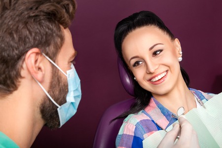 Young beautiful woman patient with white straight teeth sitting in the dental chair and smilling while male doctor with beard examining her teeth with help of dental mirror. Healthcare, medicine.の写真素材