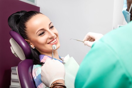 Young beautiful woman patient with white straight teeth sitting in the dental chair and smilling while male doctor with beard examining her teeth with help of dental mirror. Healthcare, medicine.の写真素材