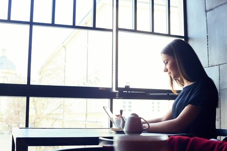 Young attractive girl sitting alone near big window in coffee shop during free time and working on tablet computer. Happy female having rest in cafe. Lifestyle, coffeeの写真素材
