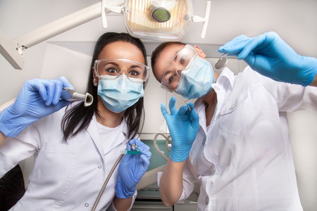 Female doctor dentist and her assistant in masks and white uniforms holding in their hands dental mirror, probe and other equipment, looking at camera and smilingの写真素材