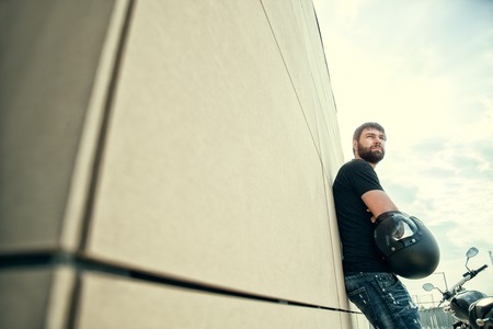 Portrait of biker men with beard in black shirt standing near a modern wall, holding helmet in his hands and looking away on sunsetの写真素材