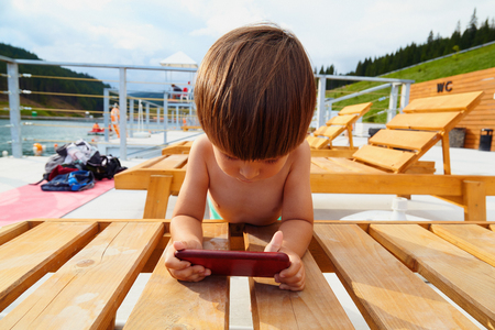 Portrait of cute little toddler boy using his smart phone, playing games on beach near mountain lake on sunny day outdoor on blue sky background.の写真素材