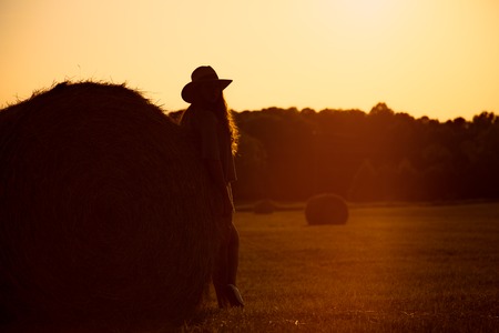 Silhouette of beautiful girl with long curly brunette hair wearing hat stand near a hay bale in summer stubble field on sunset background.の写真素材