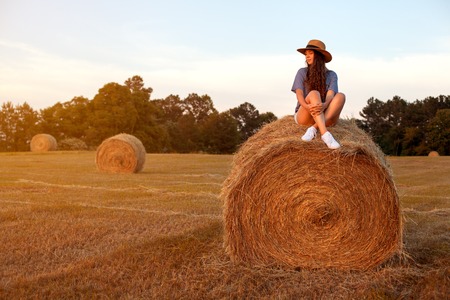 Pretty young woman with long hair in hat wearing shorts, t-shirt and white sneakers sitting on a hay bale in summer stubble field in sunset background.の写真素材