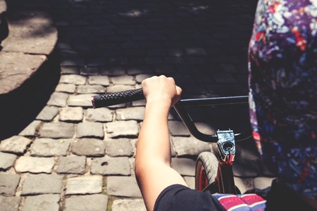 Back view of a teenage boy in a cap sitting on a ramp of a bike and holding his hands on a black handlebar of BMX bicycle in summer day on a pavement backgroungの写真素材