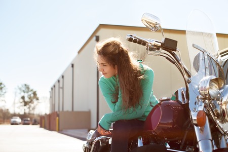 Young attractive woman with long curly hair and beautiful smile wearing green sweater and jeans is sitting on red motorcycle on summer day outdoor. Lifestyle, vacation, happiness, joy conceptの写真素材