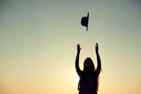 Silhouette of woman with long curly hair throwing her hat in the air at sunset outdoor.の写真素材